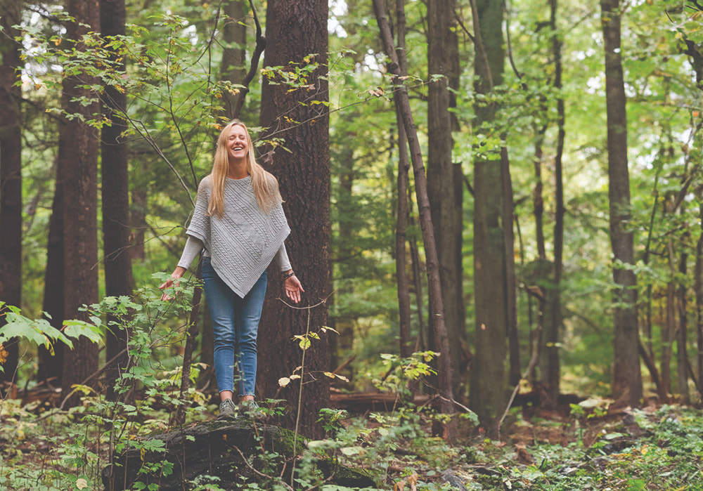 Smiling woman with arms lowered and eyes closed standing in the woods.